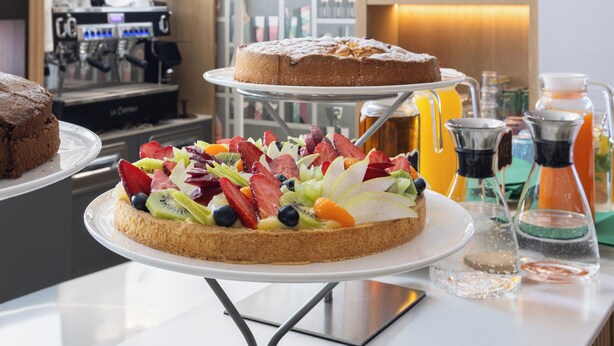 Een dessertbuffet met een taart gegarneerd met fruit, een koffiemachine en planken met flessen en glazen.