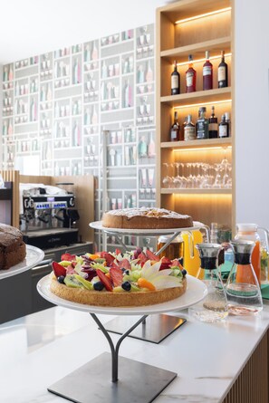A dessert station with a fruit-topped cake, a coffee machine, and shelves with bottles and glasses. - ENERGY PARK HOTEL (Vimercate)