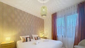 A hotel room with a large bed, a bedside table with a lamp, and a view of trees through sheer curtains. - Apt3BR,AC,WiFi, sea view, parking, 3min to beach (Fréjus)