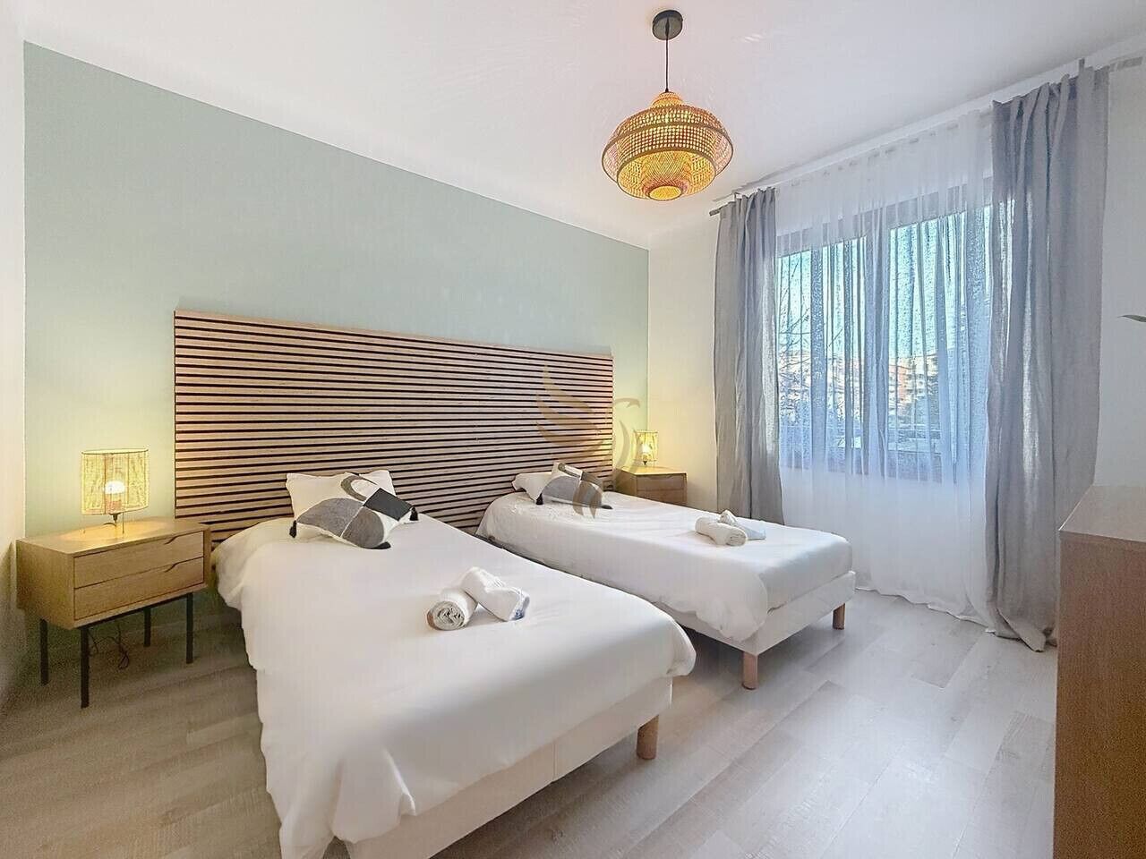 A hotel room with two beds, a large window with curtains, a bedside lamp, and a hanging light fixture.