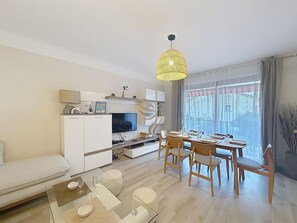 A modern living room with a sofa, dining table, chairs, a TV, and a kitchen area with cabinets and a hanging light fixture.