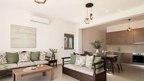 A modern living room with a sofa, coffee table, and dining area. There is a kitchen in the background with wooden cabinets and a countertop. - Stone villa with private pool close to Marathi beach (Kounoupidiana, Marathi, Chania)