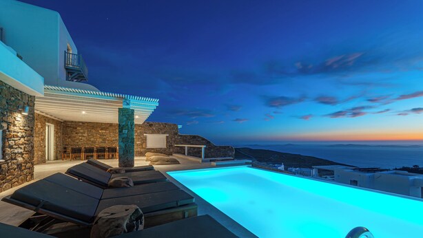 Área de piscina com espreguiçadeiras, uma parede de pedra e vista para o mar ao entardecer.