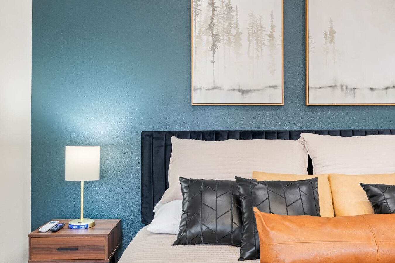 Ein Schlafzimmer mit blauer Wand, einem Bett mit vielen Kissen, einem Nachttisch mit Lampe und zwei gerahmten Bildern.