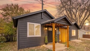A small, single-story building with a dark exterior, a front porch with string lights, and a visible mailbox. - The Creekside Pump House (Spearfish)