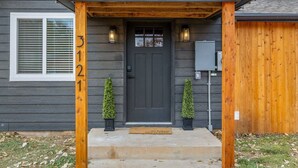 A house with a dark gray exterior, a wooden porch, a black front door, and two small potted plants on either side. - The Creekside Pump House (Spearfish)