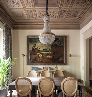 A dining area with a chandelier, a large landscape painting, wicker chairs, and a table.
