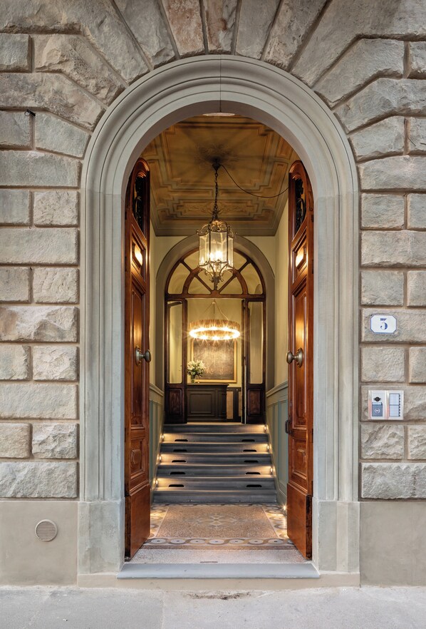 A grand entrance with a stone archway, a chandelier, and a staircase leading to a room with a large mirror and a floral arrangement. - This Time Tomorrow in Florence (Florence)