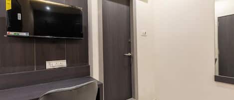 A room with a dark wood desk, a flat-screen TV mounted above it, a door with a handle, and a power outlet on the wall.