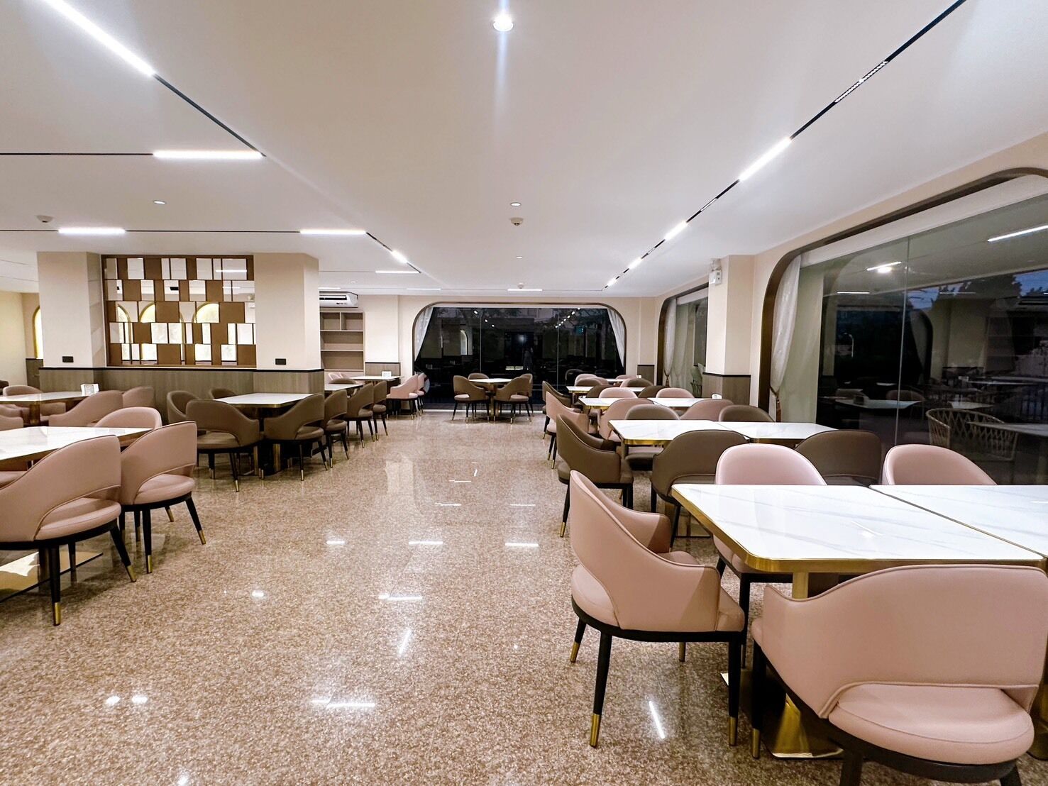 A spacious dining area with multiple tables and chairs, a modern ceiling design, and a large window providing a view outside.