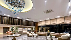 A spacious lobby with a modern design, featuring a central seating area with sofas and armchairs, a decorative chandelier, and a large window offering a view outside.