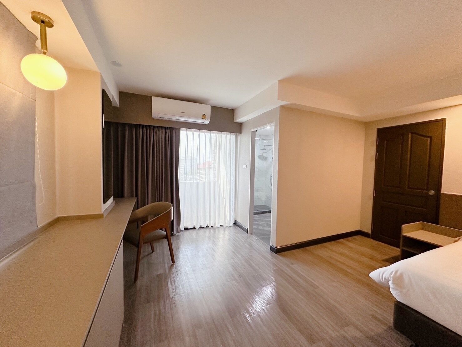 A hotel room with a bed, a desk, a chair, a window with curtains, an air conditioning unit, and a door.