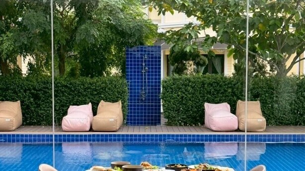 A dining area with a pool view, featuring a table with food and two chairs, surrounded by a wooden deck and a glass wall.