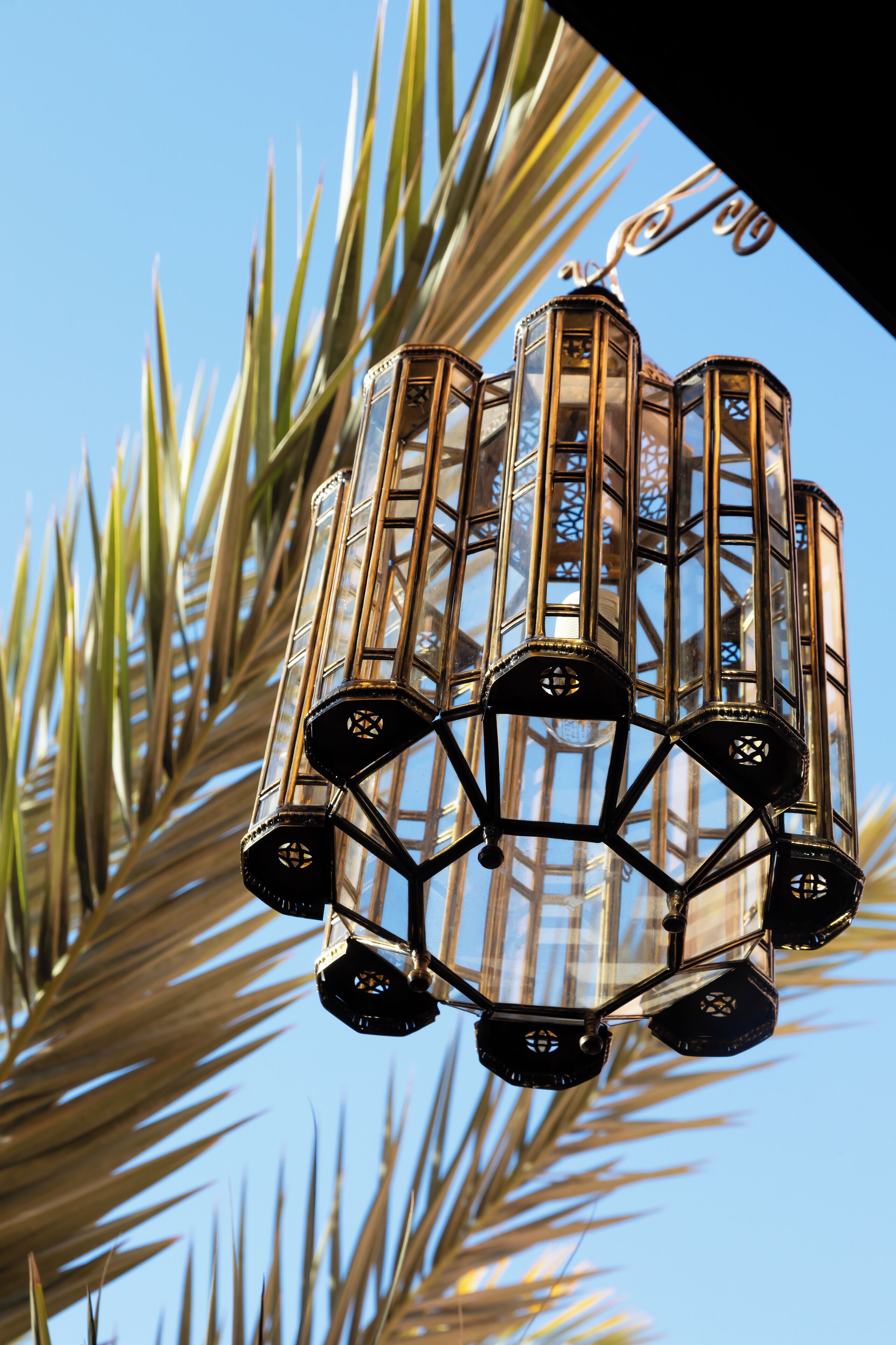 A hanging light fixture with multiple glass panels and a geometric design, set against a backdrop of palm leaves.