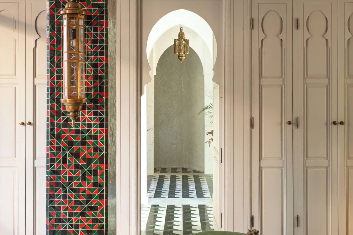 A room with a patterned column, a bed with green bedding, and a view of a tiled corridor leading to a door.