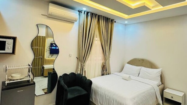 A hotel room with a bed, a chair, a desk with a lamp, a mirror, and curtains. - Studio(S8)@LEKKI PHASE 1, 24/7 Pwr by NgoZiLiving


 (Lekki)