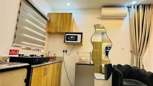 A compact kitchen with a gas stove, microwave, and a small refrigerator. There is a wooden cabinet and a mirror on the wall. - Studio(S8)@LEKKI PHASE 1, 24/7 Pwr by NgoZiLiving


 (Lekki)