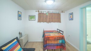 A small, well-lit room with a bed, a table, and a chair. There are framed pictures on the walls and a curtain with a pattern.