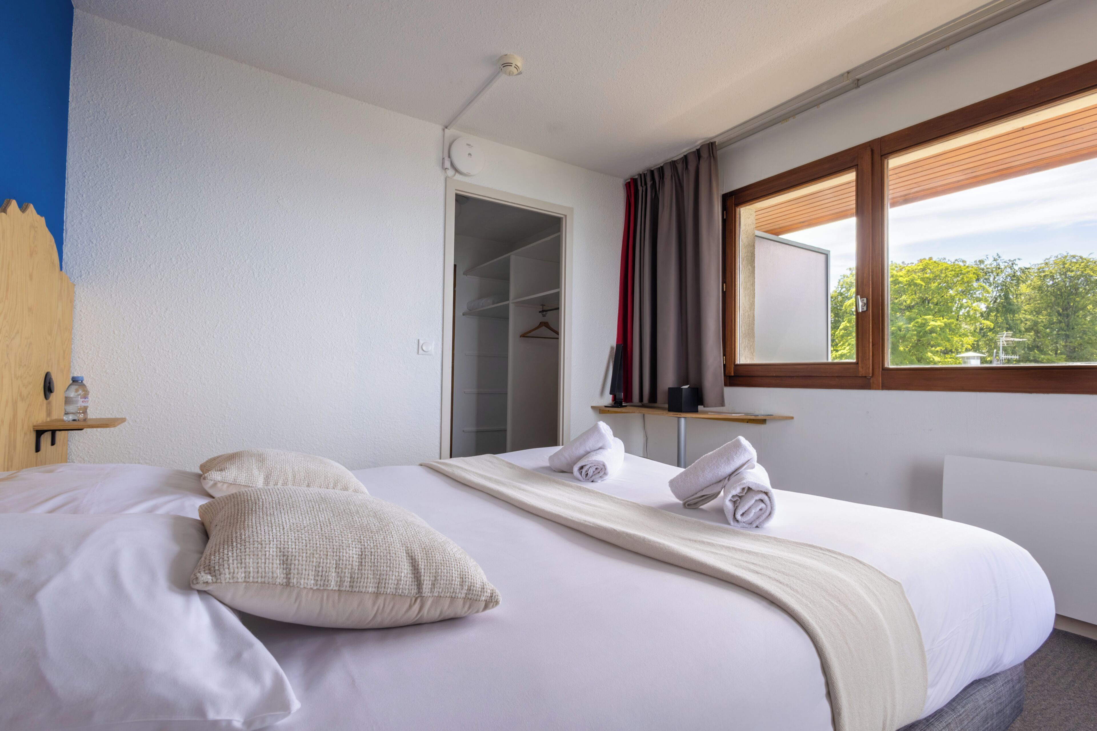 Ein ordentlich bezogenes Bett mit Kissen, Handtuch und Decke. Dazu ein Holz-Kopfteil, ein Nachttisch mit einer Flasche und ein Fenster mit Vorhängen.