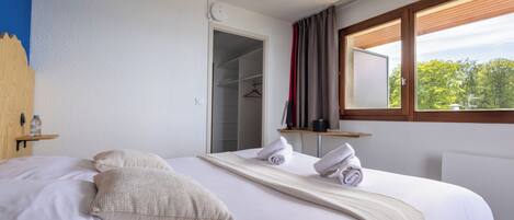 Ein ordentlich bezogenes Bett mit Kissen, Handtuch und Decke. Dazu ein Holz-Kopfteil, ein Nachttisch mit einer Flasche und ein Fenster mit Vorhängen.
