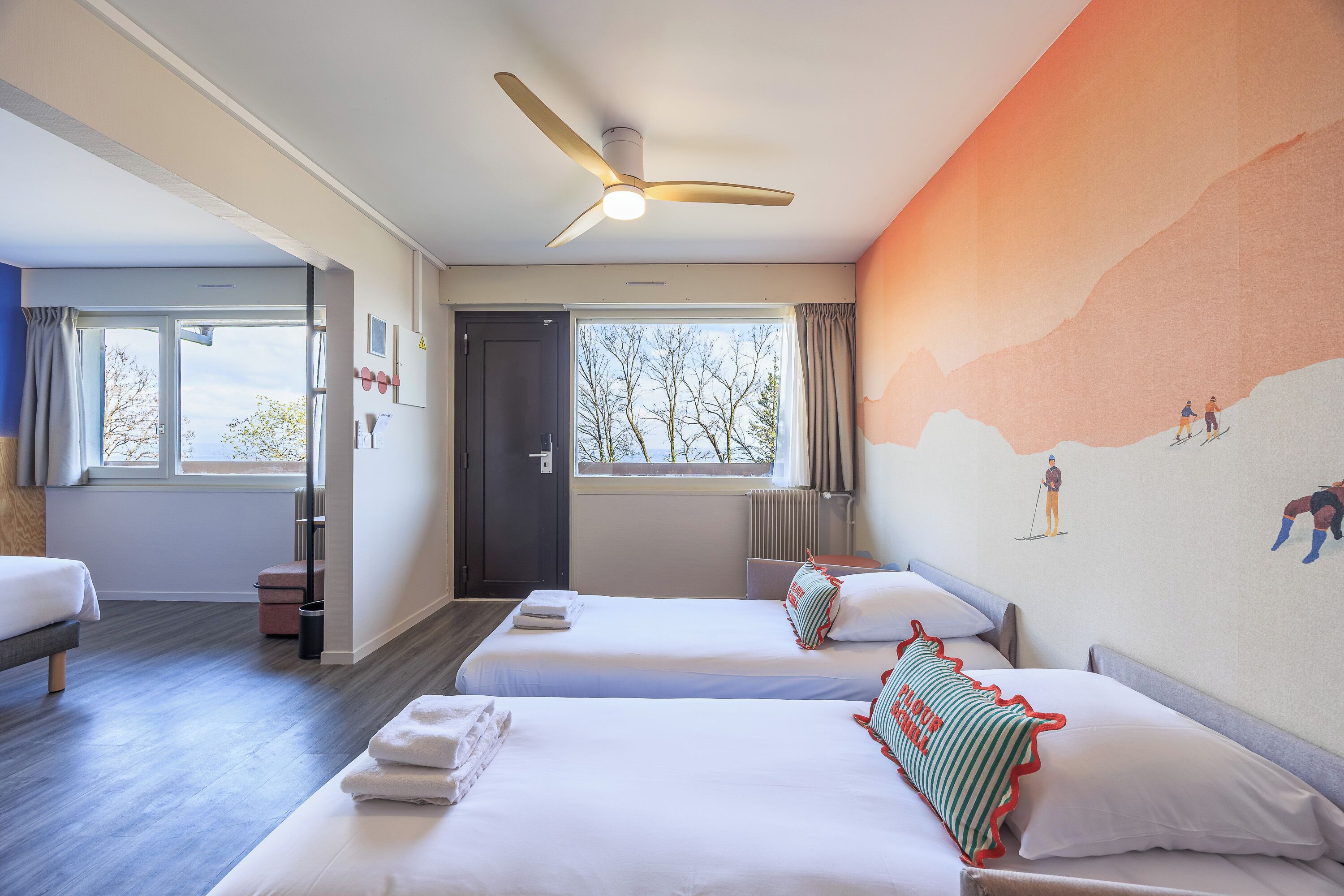 A hotel room with two beds, a ceiling fan, a window with a view, and a mural on the wall.