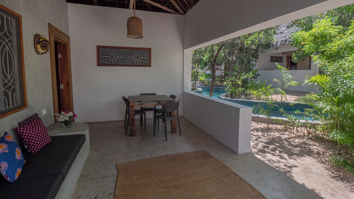 A room with a dining table, chairs, a sofa with cushions, a hanging light fixture, a decorative wall piece, and a view of an outdoor swimming pool surrounded by greenery.