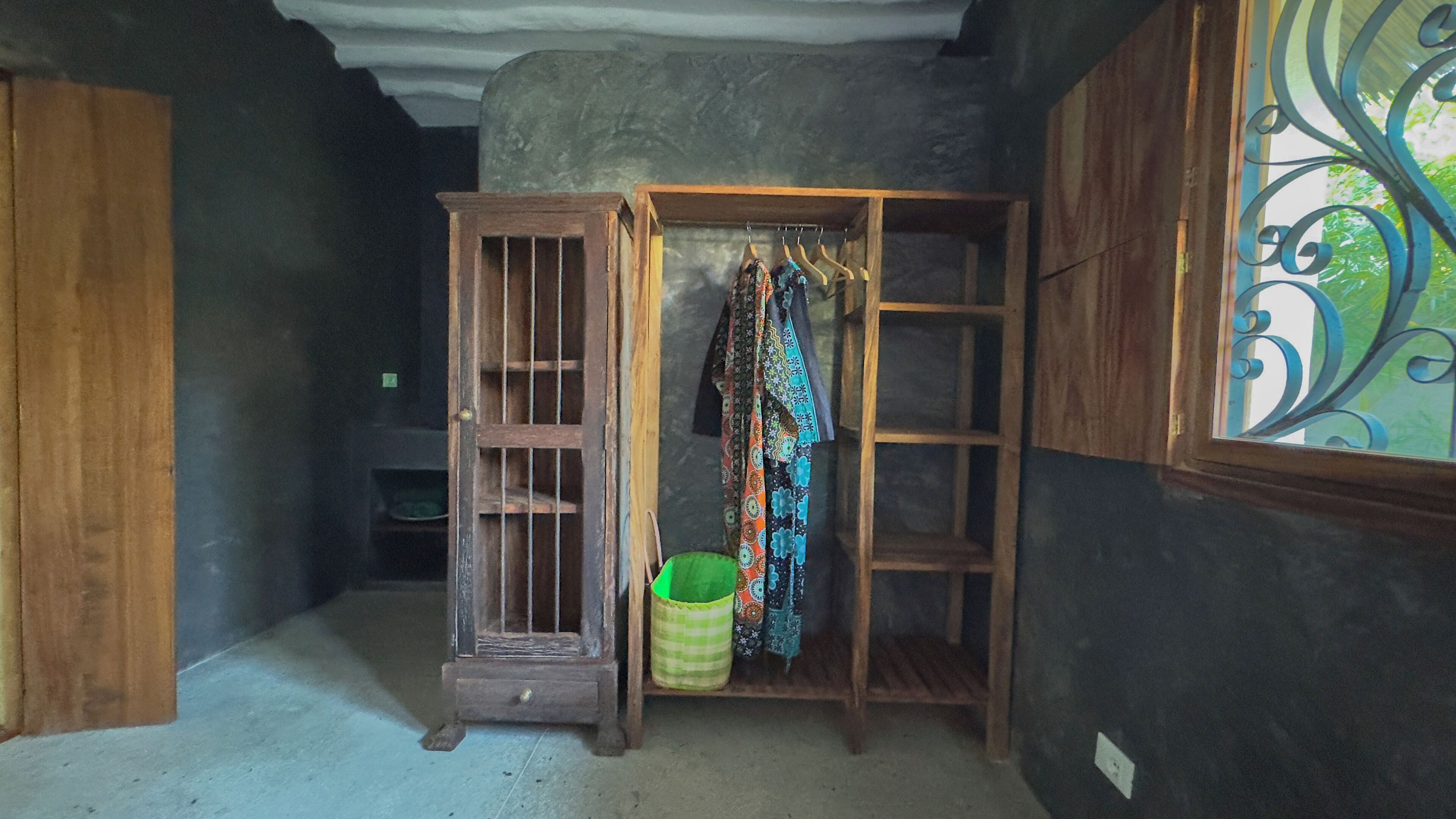 A room with a wooden wardrobe, a hanging rack with colorful fabrics, and a green trash bin.