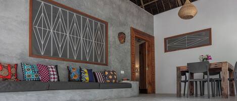 Una habitación con un sofá gris, una mesa de comedor de madera con sillas, una lámpara colgante y un ventanal grande con diseños geométricos.