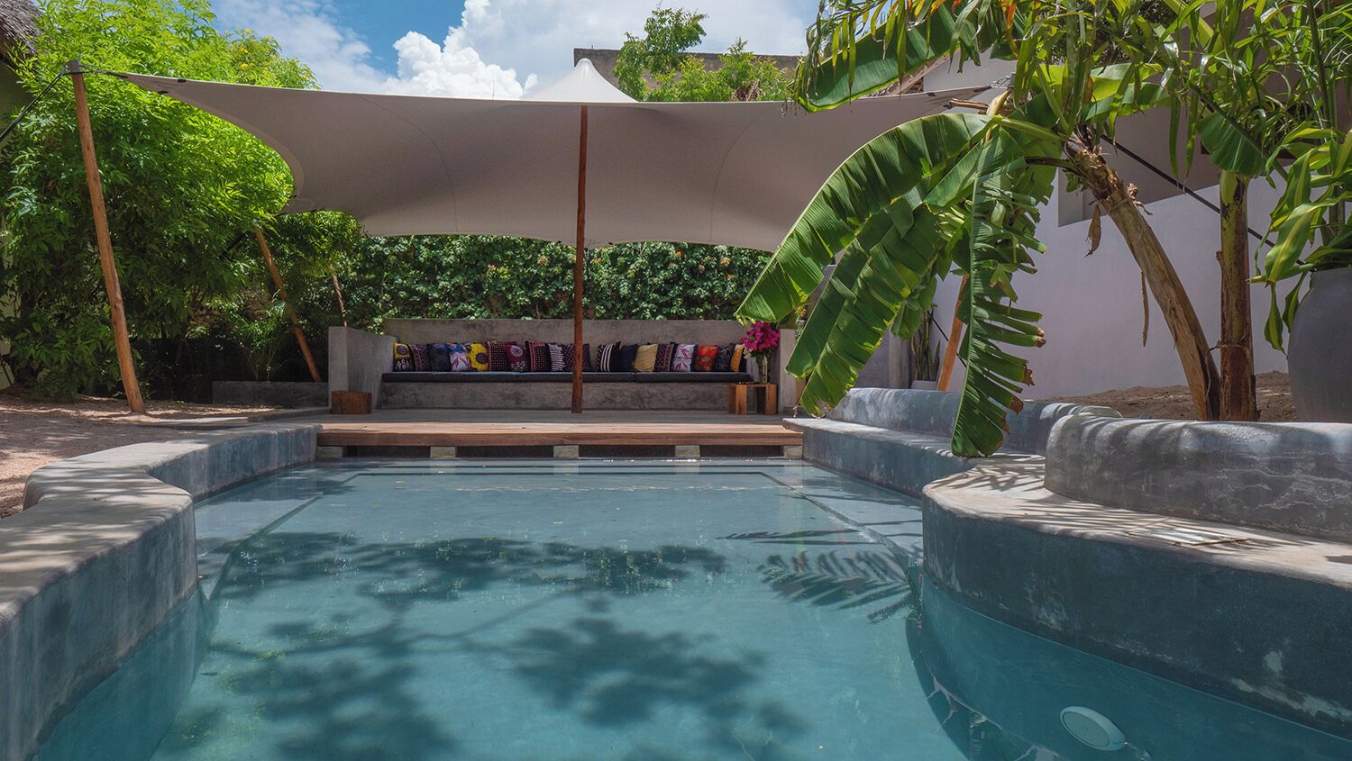 A swimming pool with a built-in bench area, surrounded by stone walls and tropical plants.