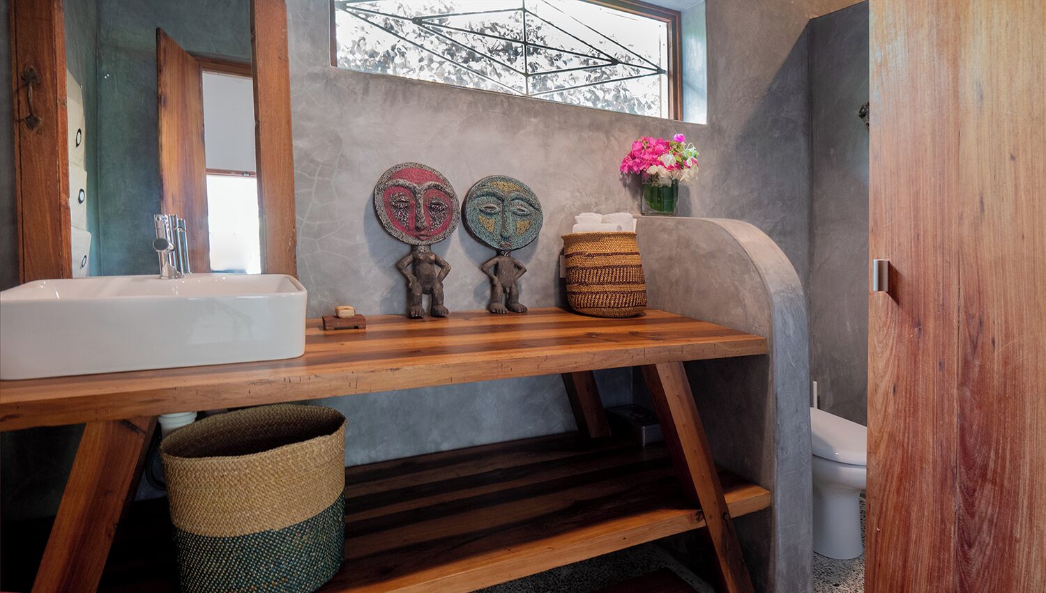 A bathroom with a wooden vanity, a sink, two decorative masks, a woven basket, and a window with a view of trees.