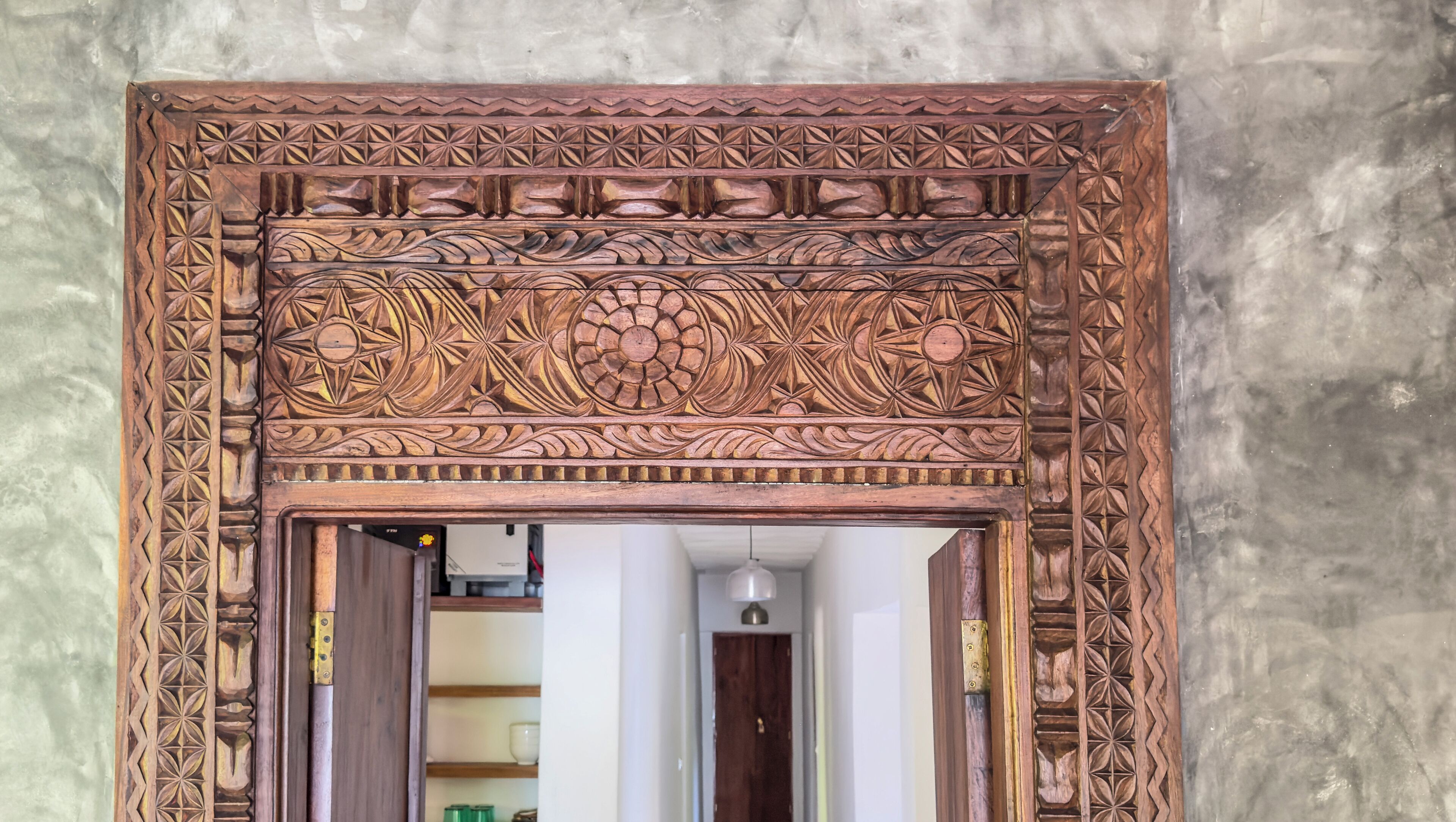 A wooden carved doorway with intricate designs, leading into a room with a white door and a shelf with items.