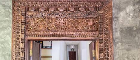 A wooden carved doorway with intricate designs, leading into a room with a white door and a shelf with items.