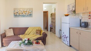 A living room with a sofa, a coffee table with flowers, a kitchen area with a refrigerator and a kettle, and a hallway leading to another room.