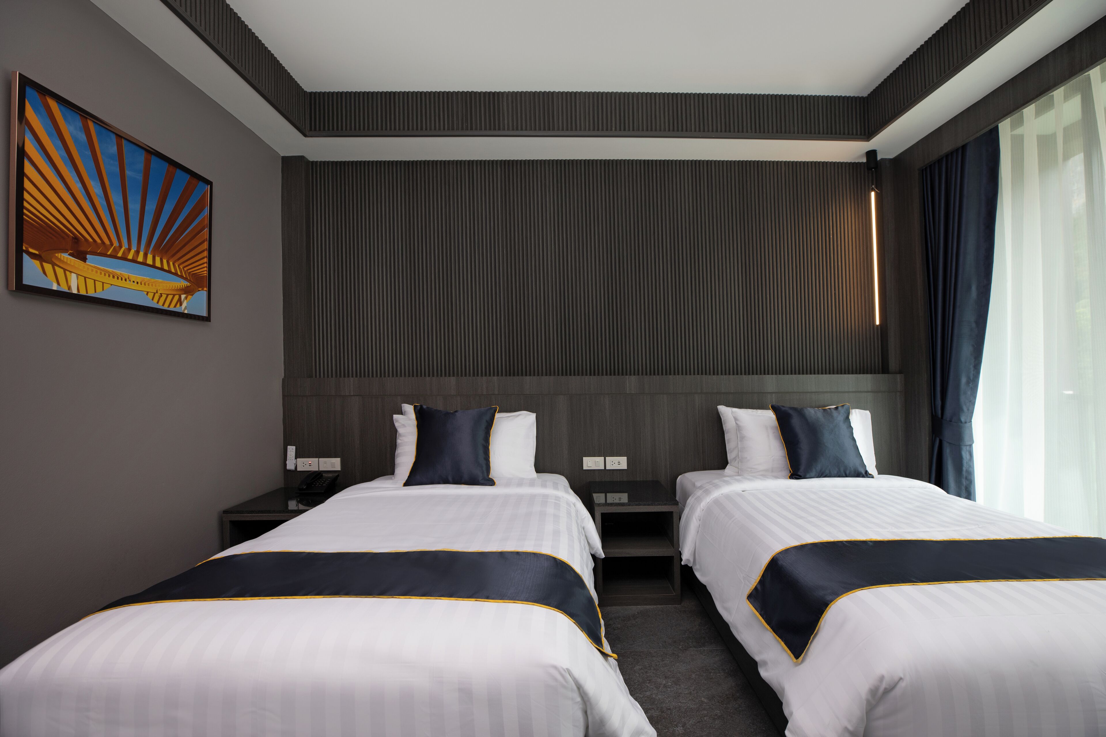 A hotel room with two beds, a large window with curtains, a wall-mounted artwork, and a nightstand with a phone.