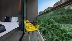 Ein modernes Hotelzimmer mit einem großen Bett, einem kleinen Tisch und einem gelben Stuhl. Das Zimmer verfügt über ein großes Fenster mit Blick auf üppige Vegetation und Berge.