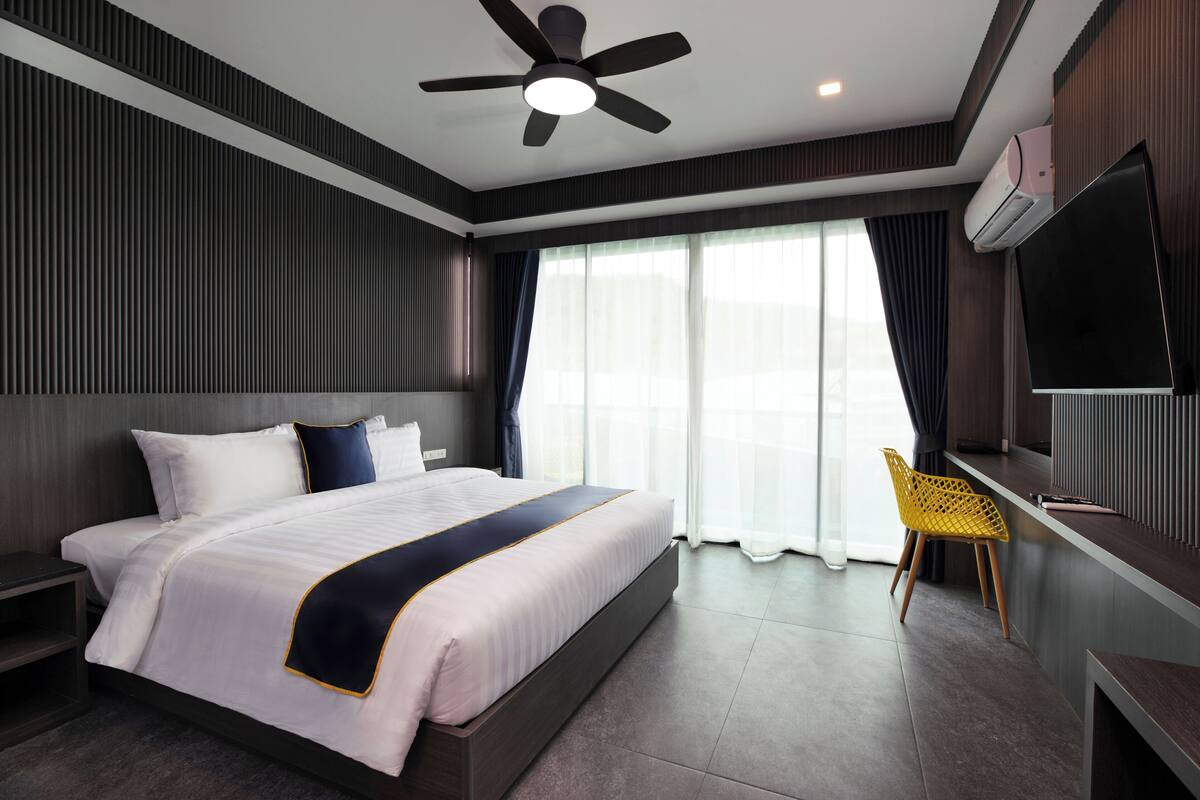 Habitación de hotel moderna con una cama grande, ventilador de techo, televisor de pantalla plana y una zona de estar con una silla.
