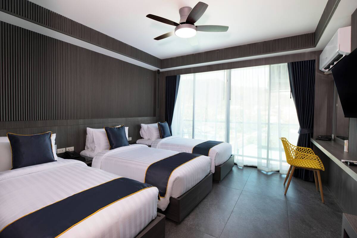 Habitación de hotel con dos camas, un ventilador de techo, un ventanal con cortinas y una silla amarilla.