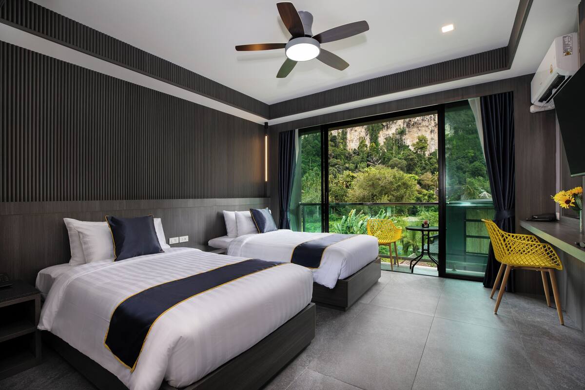 Una habitación de hotel moderna con dos camas, un ventilador de techo, un ventanal con vistas a zonas verdes y una mesita con sillas amarillas.