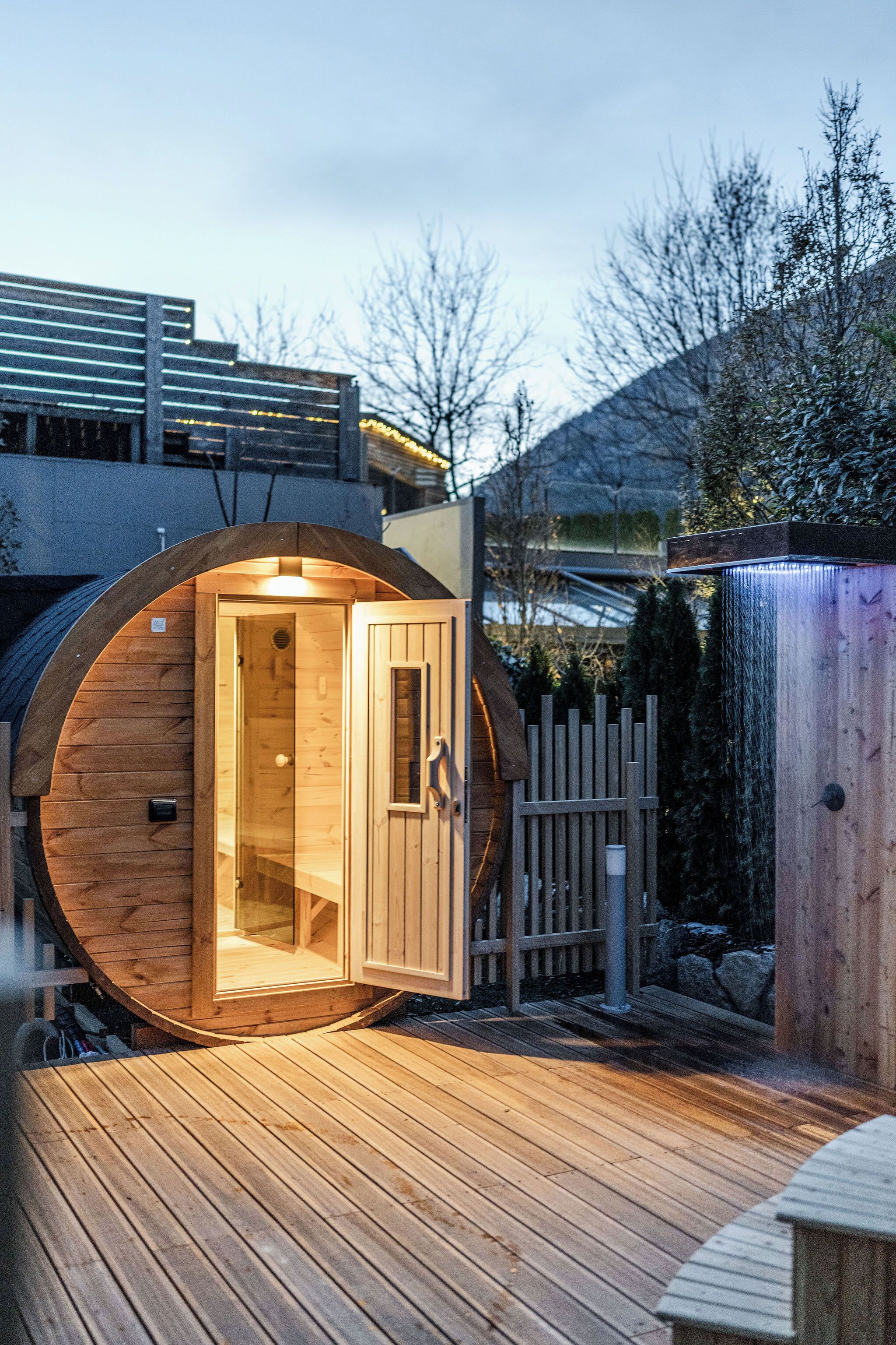 A wooden sauna structure with a door and a small window, surrounded by a wooden deck and a fence.