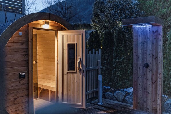 A wooden sauna with a glass door and a bench inside, surrounded by a wooden fence and trees. - Hotel Garni Meledrio (Dimaro Folgarida)