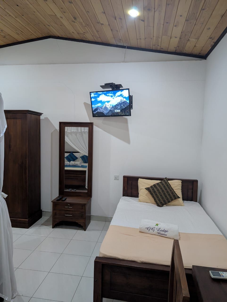 A bedroom with a bed, a TV mounted on the wall, a wooden wardrobe, and a bedside table.