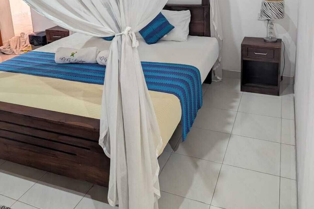 A bedroom with a bed under a white canopy, a bedside table with a lamp, and a patterned rug on the floor.