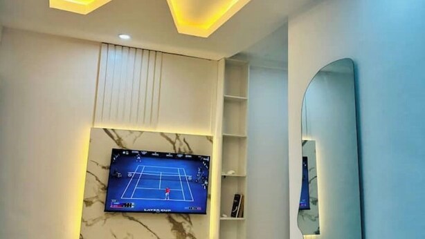 A room with a flat-screen TV mounted on a wall, a ceiling with a zigzag light design, and a large mirror on the right wall. - Studio(S3)@LEKKI PHASE 1, 24/7 Pwr by NgoZiLiving

 (Lekki)