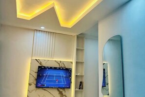 Une pièce dotée d’un téléviseur à écran plat fixé au mur, d’un plafond orné d’un motif lumineux en zigzag et d’un grand miroir sur le mur de droite.