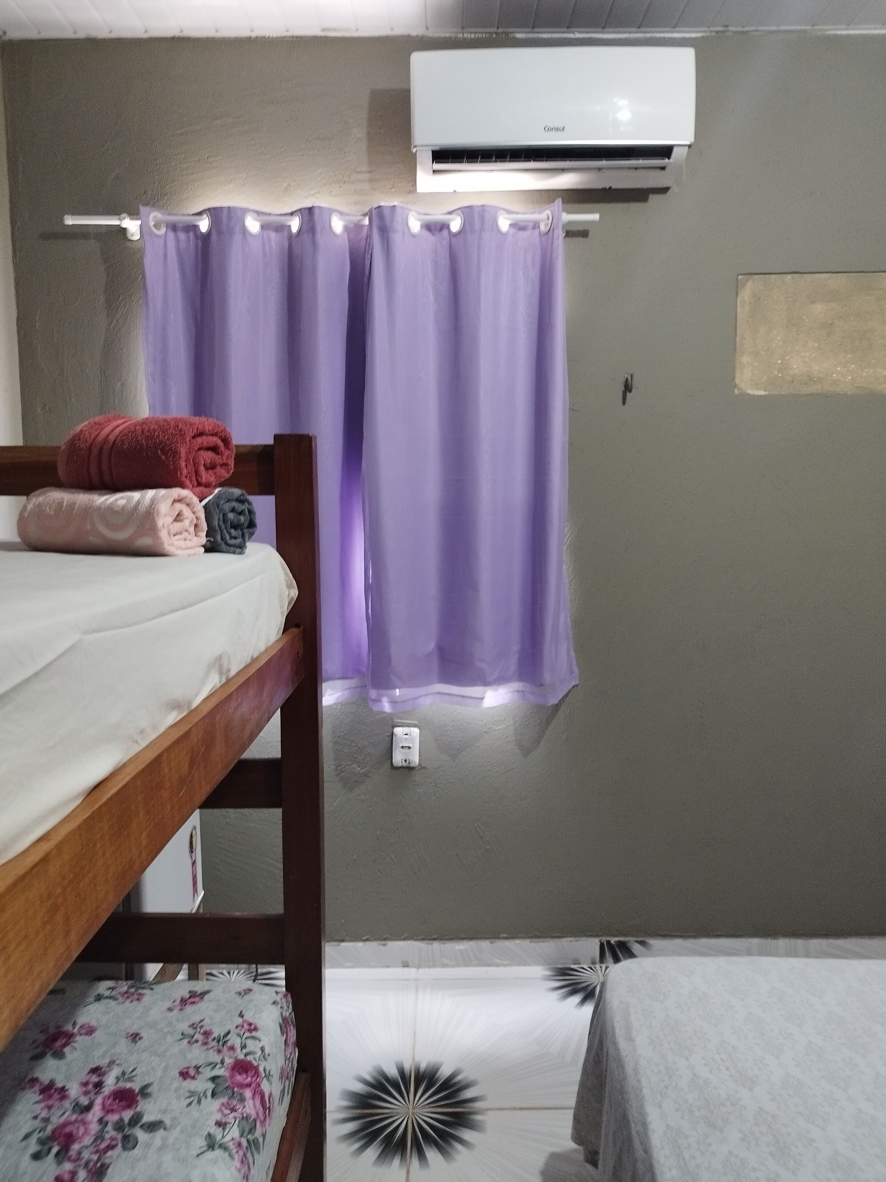 Una habitación con una cama litera, una cama individual, cortinas moradas, aire acondicionado y un enchufe en la pared.