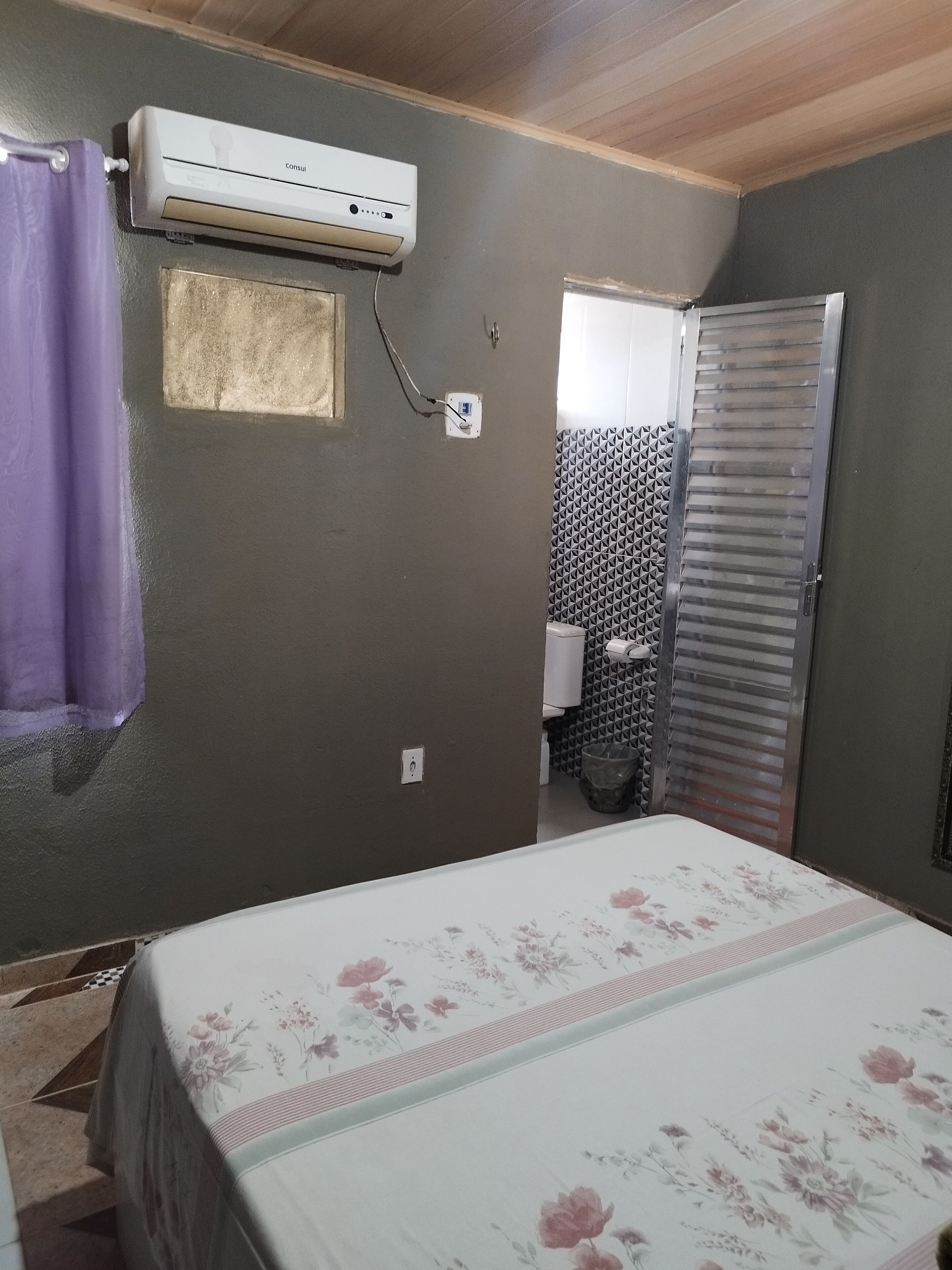A bedroom with a bed, an air conditioner, a bathroom with a toilet, and a shower area.