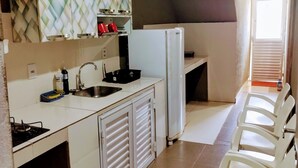 A compact kitchen with a sink, stove, and white cabinets. There are two white chairs and a window with blinds. - Pousada Temporada no Atalaia (Salinópolis)
