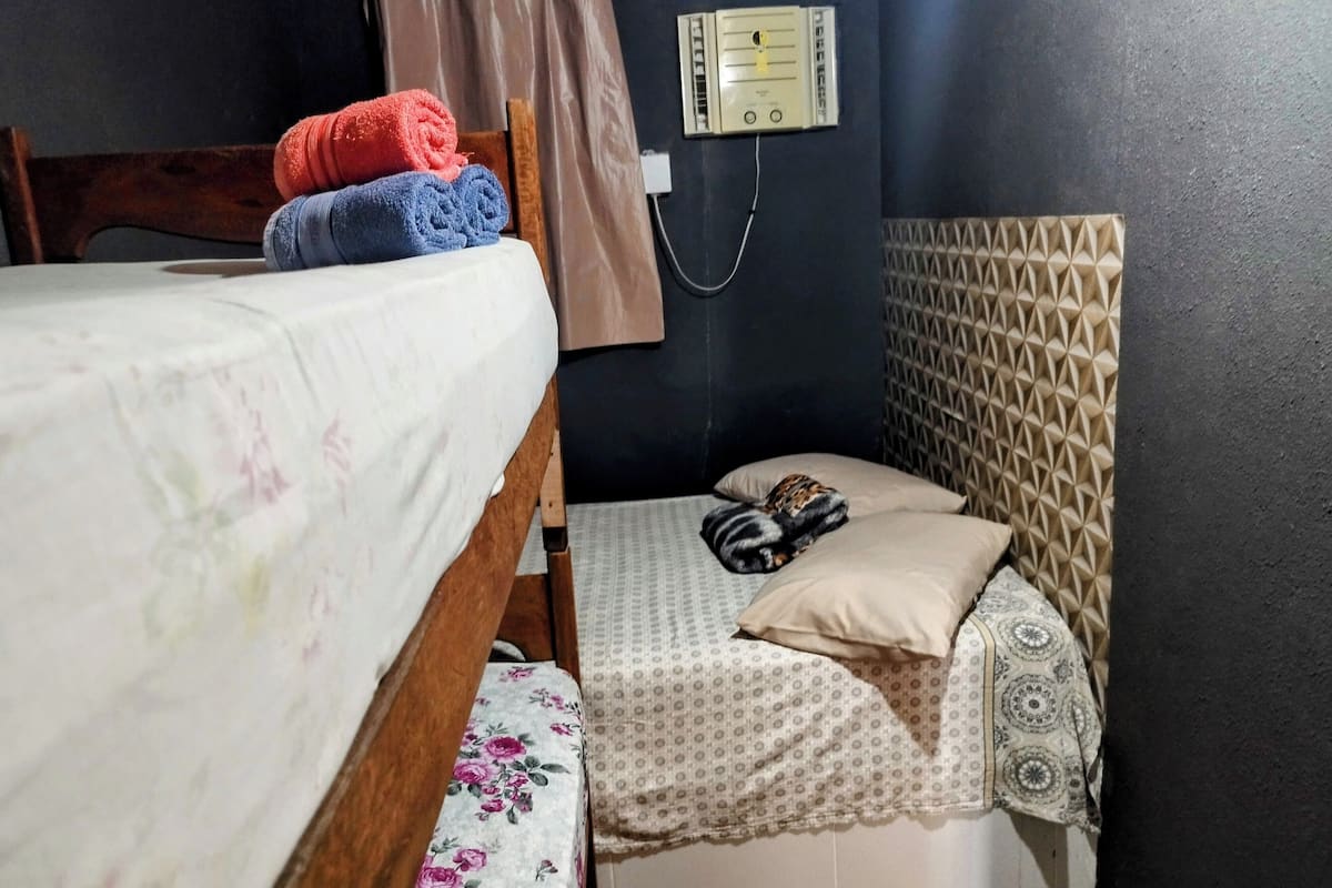 Pieni, hämärästi valaistu huone, jossa on kaksi sänkyä. Toisessa on kukallinen päiväpeite ja toisessa kuviollinen päiväpeite. Seinässä on pistorasia ja kuviollinen seinäpaneeli.