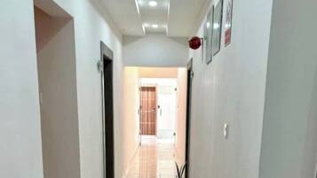 Un couloir avec un sol carrelé, une porte à gauche et un mur sur lequel se trouvent un interrupteur et une petite plante.