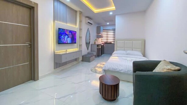 Ein modernes Hotelzimmer mit einem großen Bett, einem an der Wand befestigten Fernseher, einer kleinen Sitzecke und einem im Hintergrund sichtbaren Badezimmer.